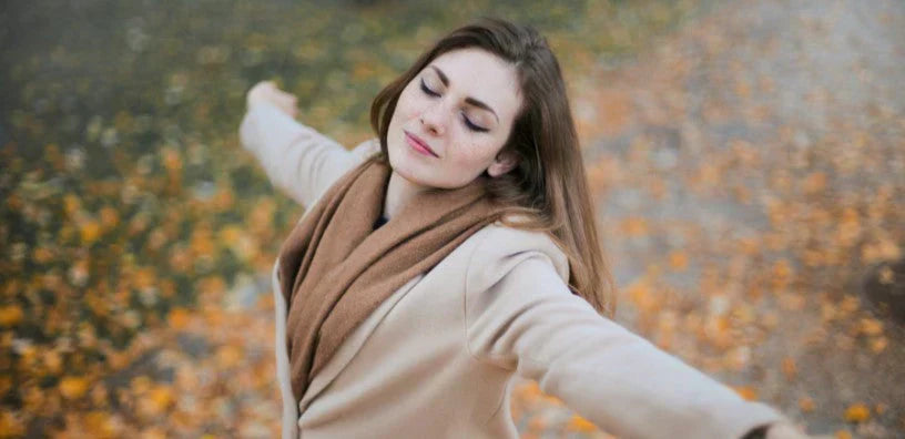 A woman wearing a beige coat and brown scarf stands outdoors with her arms outstretched and eyes closed, enjoying a peaceful autumn day surrounded by fallen leaves.