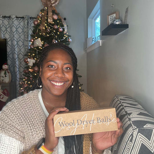 Smiling woman holding a box of wool dryer balls, seated on a couch with a decorated Christmas tree in the background.