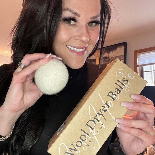 Smiling woman holding a wool dryer ball and a box labeled “Wool Dryer Balls – All Natural,” standing in a cozy, well-lit room.
