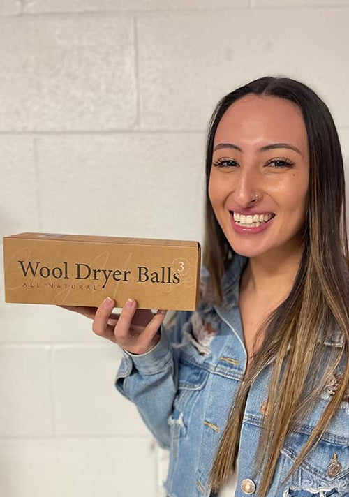 Smiling woman in a denim jacket holding a box of all-natural wool dryer balls against a white brick wall.