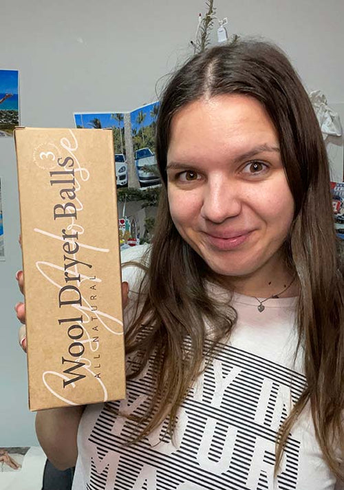 Smiling woman holding a box of all-natural wool dryer balls, standing indoors with travel photos on the wall behind her.