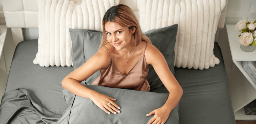 Young woman sitting on a bed with dark grey bamboo sheets and pillowcases, highlighting luxury bamboo bedding that’s breathable, soft, and cooling.