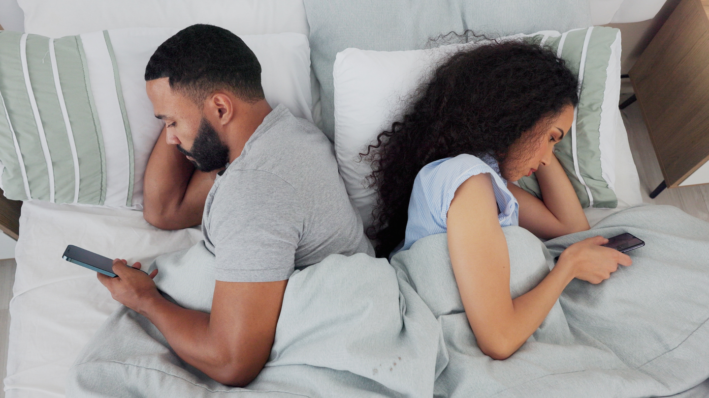 Young couple in bed, turned away from each other, staying up to scroll on their cell phones