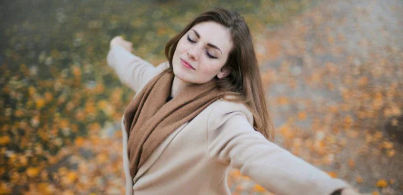 A woman wearing a beige coat and brown scarf stands outdoors with her arms outstretched and eyes closed, enjoying a peaceful autumn day surrounded by fallen leaves.