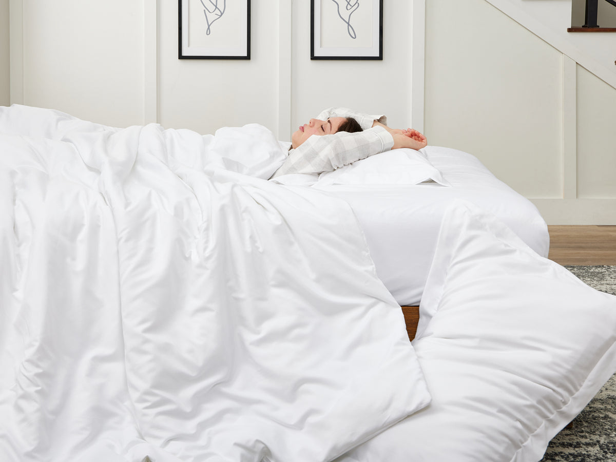 Woman lying in bed under a fluffy white comforter, stretching with eyes closed in a cozy, well-lit bedroom.