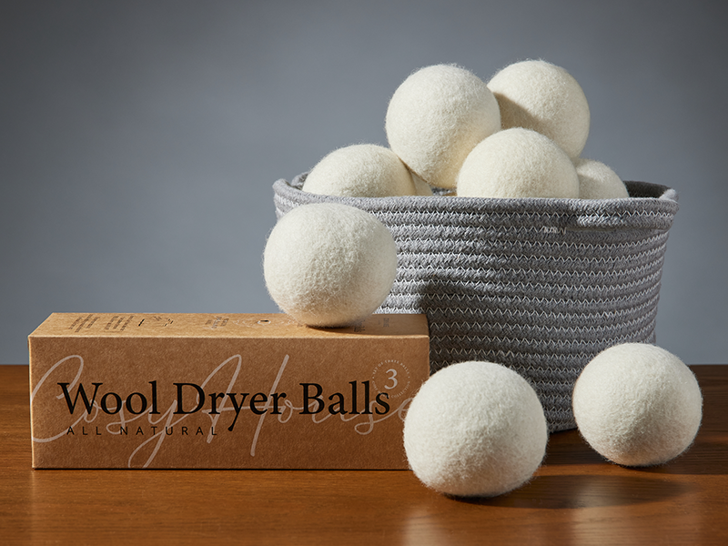 Box of wool dryer balls with several white balls displayed in front and inside a gray woven basket on a wooden surface.