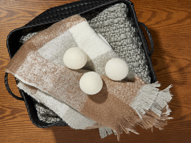 Three wool dryer balls on a folded neutral-toned blanket inside a black woven basket, set on a wooden surface.