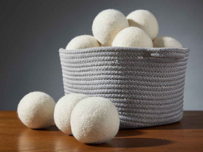 Wool dryer balls in and around a gray woven basket, displayed on a wooden surface with a neutral background.