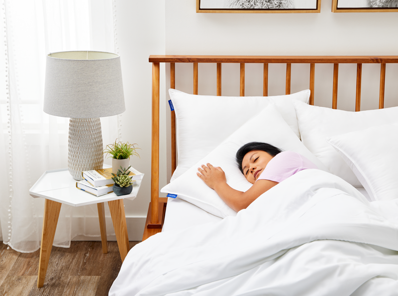 Woman sleeping peacefully on her side in a white bed with fluffy pillows, next to a wooden headboard and a modern white nightstand holding a textured lamp, books, and small potted plants.