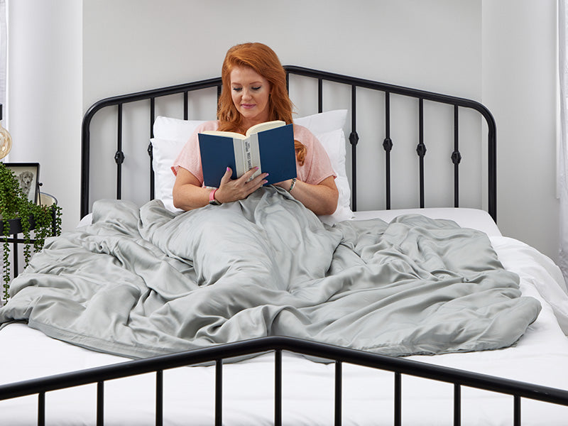 Woman sitting up in bed, reading a book while wrapped in a gray blanket in a cozy, minimalist bedroom.